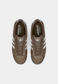 Ténis de camurça castanha com riscas brancas, bico redondo e atacadores planos. Marca notável na língua e na sola interior.