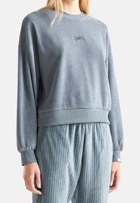 Kvinna som bär en ljusblå velourtröja med lång ärm, ribbade ärmslut och broderad logotyp, ihop med lösa ribbade blå byxor.