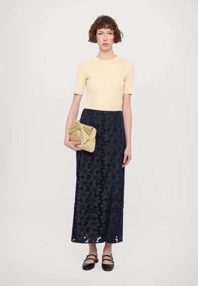 Lys gul kortermet topp paret med et marineblått maxiskjørt i blonder med blomsterdesign, holder en gullfarget, teksturert clutch-veske. Brune sko.