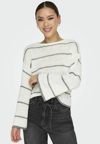 Pull en tricot blanc avec des rayures grises, présentant un grand col et des manches à cloche, associé à un jean sombre taille haute.