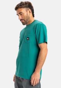 Tyrkysové tričko s krátkým rukávem vyrobené z bavlny s kulatým výstřihem, s náprsní kapsou na levé straně a černou nášivkou loga. Hladká textura.