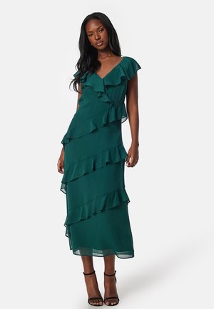 Robe maxi en teal à volants avec un décolleté en V, des manches courtes et des couches superposées. Tissu doux et fluide avec un design légèrement transparent. Sandales à talons noires à lanières.