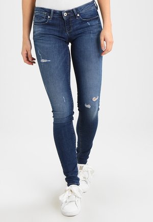 Schmal geschnittene blaue Jeans mit kleinen Rissen, getragen mit weißen Sneakern mit übergroßen Schleifen-Schnürsenkeln auf hellem Hintergrund.