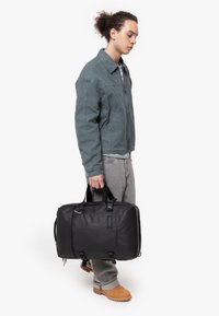 Borsa a duffle nera con due manici superiori, materiale testurizzato, chiusura con zip e spallacci regolabili. Modello che indossa una giacca teal e pantaloni grigi.