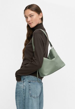 Woman with long brown hair wearing a dark turtleneck and blue jeans carries a muted green shoulder bag, looking sideways against a plain background.