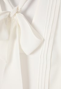Biała bluzka z wiązanym kołnierzykiem w kształcie kokardy, pionowymi plisami i gładką fakturą materiału. Akcenty z delikatnym drapowaniem wokół dekoltu.