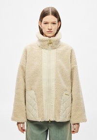 Helebeežist beeži fliisjakk kõrge kraega, millel on tikitud kreemjad taskud, kuldse tõmblukuga riistvara ja lõdvestunud varrukad.