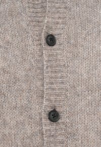 Cardigan bej deschis, realizat dintr-un material tricotat texturat. Prezintă dungi verticale și nasturi negri pe placa frontală.