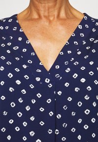 Top de tecido azul-marinho com decote em V, apresentando um padrão geométrico branco de quadrados e diamantes. Textura suave, linhas limpas e sem hardware visível.