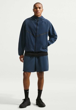 Ung mand stående iført en mørkeblå lynlåstrøje, matchende shorts, sorte sokker og sorte sportssko på en ensartet lys baggrund.