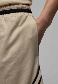 Pantalones cortos de atletismo beige con una superficie texturizada que presenta pequeñas perforaciones, una cinturilla elástica negra y bolsillos laterales. Acentos a rayas negras.