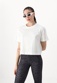 Bela skrajšana bombažna majica s kratkimi rokavi, kombinirana z črnimi leginami z leopardjim vzorcem. Model nosi bele sončne očala. Minimalistični detajl logotipa.