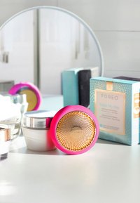 Appareil FOREO en rose vif avec une surface texturée dorée, placé près de pots blancs et argentés, ainsi qu'une boîte bleue avec des accents décoratifs.
