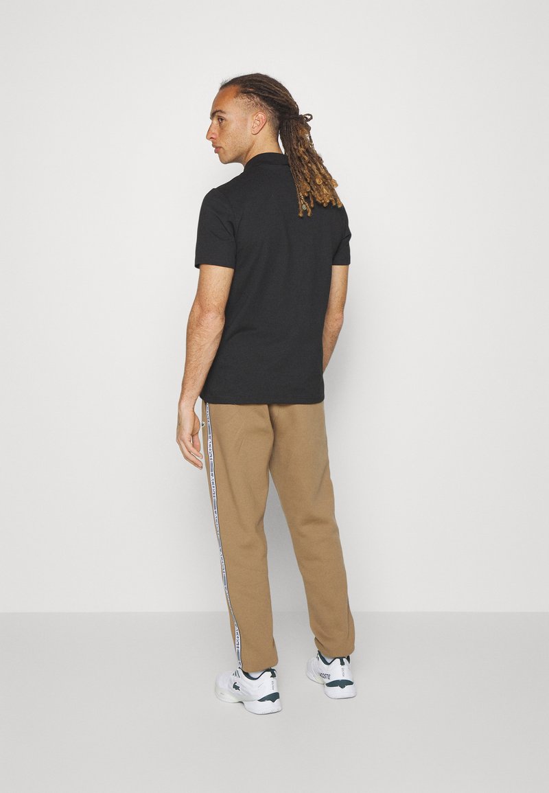 Polo noir à manches courtes associé à un pantalon beige avec des détails de rayures sur les côtés. Le modèle se tient avec une expression neutre, dos à la caméra.