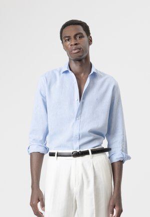 Hombre con camisa azul claro abotonada con las mangas remangadas y pantalones blancos con cinturón negro, de pie frente a un fondo claro y liso.