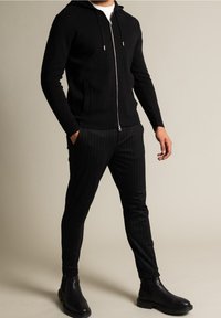 Sudadera negra con cremallera y textura suave, adornada con detalles en plata, combinada con pantalones de rayas verticales negros y botas de tobillo negras.