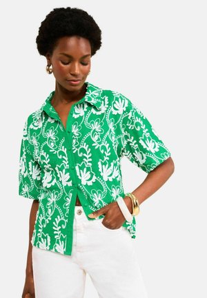 Frau in einem grünen kurzärmeligen Hemd mit weißen Blumenmustern und weißen Hosen, trägt goldene und weiße Armbänder, schaut nach unten.