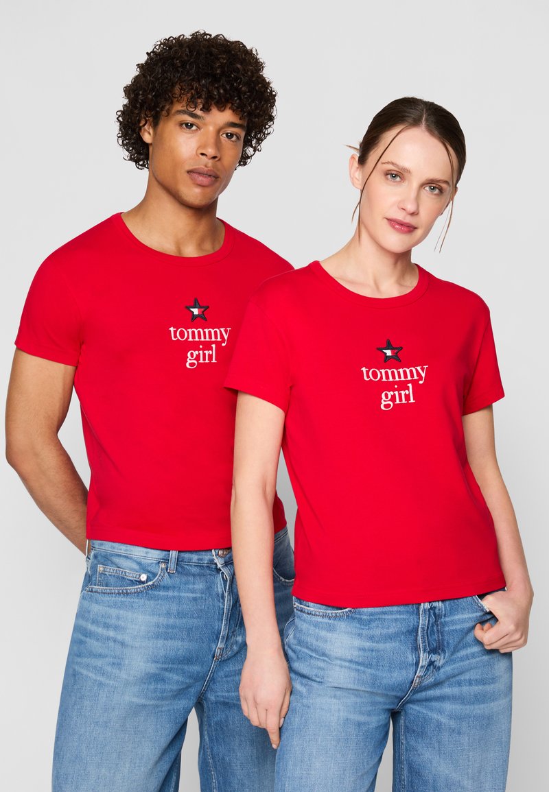Raudoni medvilniniai marškiniai su trumpomis rankovėmis, su balta "tommy girl" tekstu ir žvaigždutės grafika. Dėvimi su mėlynomis džinsinėmis kelnių.