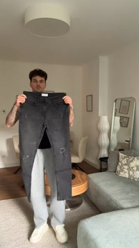 Jeans in denim nero con caratteristiche destrutturate, tenuti in mano contro uno sfondo neutro. Il materiale sembra morbido con una finitura leggermente scolorita.