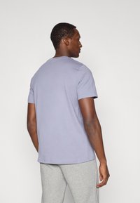 Κοντομάνικο, ανοιχτό μωβ μπλουζάκι από απαλό ύφασμα, στρογγυλή λαιμόκοψη, ελαφρώς χαλαρή εφαρμογή, φορεμένο με ανοιχτό γκρι παντελόνι φόρμας, πίσω όψη.