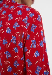 Tissu rouge avec un motif répétitif de formes abstraites bleues et blanches, présentant une texture lisse et un léger éclat.