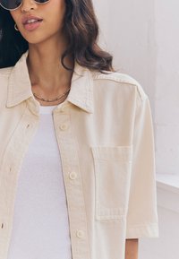 Camicia beige con bottoni e tasca applicata, realizzata in tessuto leggero. Indossata sopra una canotta bianca a coste, abbinata a occhiali da sole.