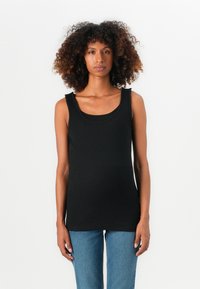 Kvinne med krøllete hår iført en svart ermeløs topp og blå jeans foran en enkel hvit bakgrunn, som vender fremover med et nøytralt uttrykk.