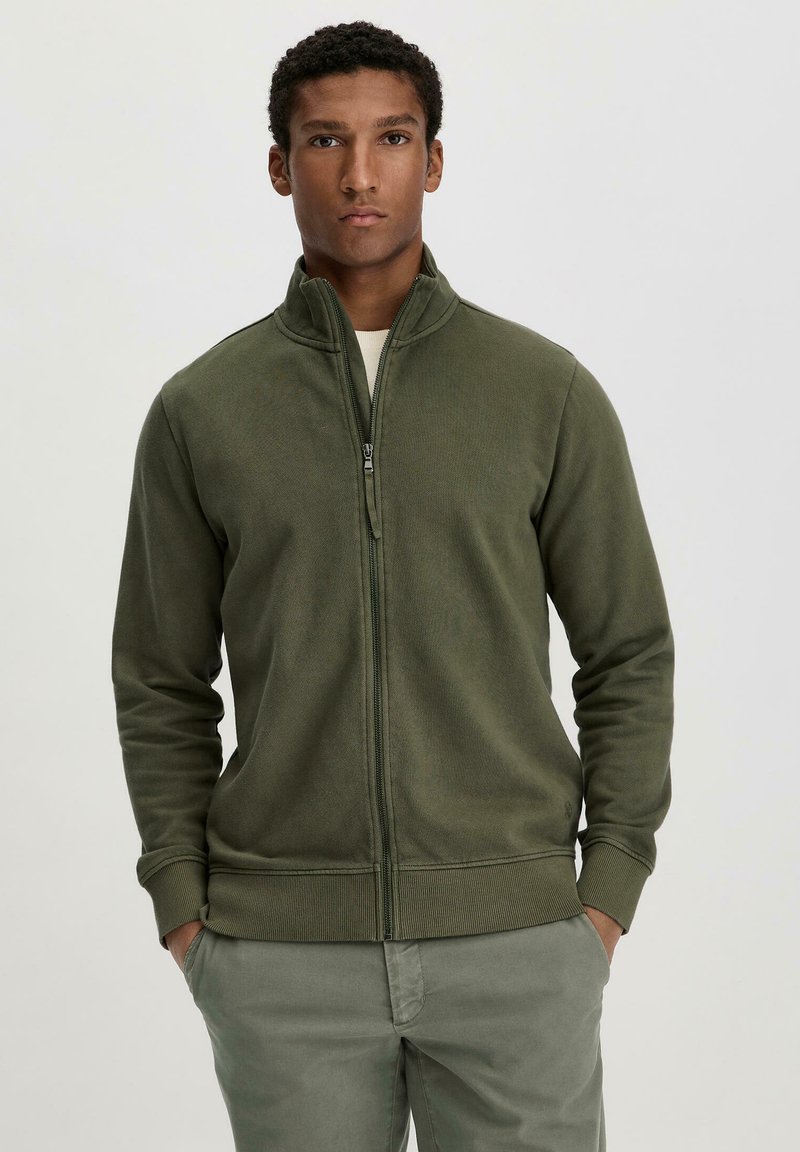 Olive green zip-up jacket in soft fabric with a high collar and ribbed cuffs. Model wears it with hands in pockets, paired with light green pants.
