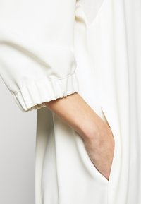 Vit långärmad plagg med sammanknutna manschetter och sidofickor; slät textur och mjukt tyg som betonar en minimalistisk design.