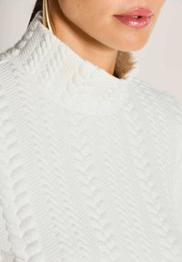 Weißer Strickpullover mit hohem, strukturiertem Kragen und erhabenen, herzförmigen Mustern auf dem Stoff, die Weichheit und Wärme betonen.