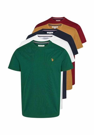 Homme debout portant un t-shirt vert à manches courtes, pantalon noir et baskets blanches sur fond clair uni.