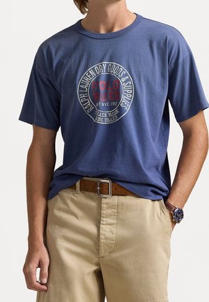 T-shirt en coton bleu marine avec un logo graphique circulaire rouge et blanc contenant du texte. Manches courtes et encolure ronde classique.