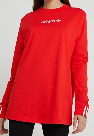 T-shirt rouge manches longues Adidas avec logo blanc sur la poitrine et trois bandes blanches aux poignets, porté par une personne portant un pantalon noir.