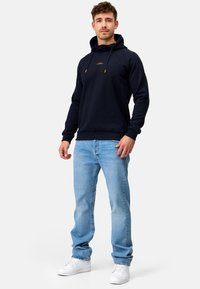 Marineblå hættebluse med snøre, med et frontlogo og ribbet kant, parret med lyseblå jeans og hvide sneakers.