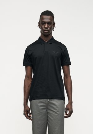 Homme debout de face portant un polo noir avec le logo "Porsche BOSS" et un pantalon gris clair, sur un fond blanc uni.