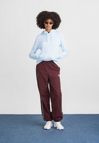 Svetlo modrá mikina s kapucňou a predným vreckom, bielym logom, v kombinácii s bordovými športovými nohavicami s modrými bočnými pruhmi a bielymi teniskami.