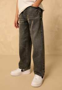 Fekete denim bővített nadrág, homogén elhalványulásos mintázattal, fehér sportcipőkkel, fekete részletekkel, semleges háttér előtt.