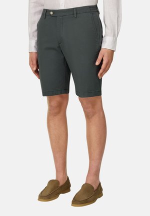 Mężczyzna w ciemnoszarych spodenkach do kolan, białej koszuli z długim rękawem i brązowych butach wsuwanych na prostym tle.