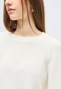 Jeune femme aux longs cheveux bruns portant un pull blanc texturé et une unique boucle d'oreille pendante en perle, sur un fond uni.