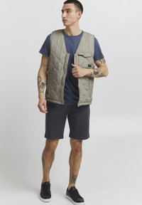 Gray quilted vest with a zip front, featuring a chest pocket, worn over a blue shirt and black shorts. Black shoes complete the outfit.