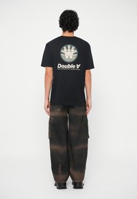 Pessoa com cabelo encaracolado está de costas, vestindo uma t-shirt preta com logótipo e texto, combinada com calças cargo largas e escuras e sapatos pretos.