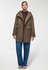 Cappotto marrone con cappuccio, chiusura a zip, grandi tasche e vestibilità comoda, abbinato a jeans a gamba larga blu e scarpe di colore chiaro.