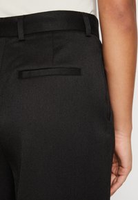 Zadní pohled na osobu v černých tailored kalhotách s jednou kapesními kapsami vzadu a poutky na opasek, ruka spočívá na boku.