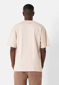 Beige bomull t-shirt med korta ärmar, med en liten orange logotyp vid nacken bak. Mjuk textur, avspänt passform och enkel design.