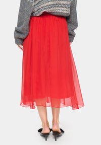 Falda larga de chiffon roja con cintura elástica, diseño en capas, textura translúcida y silueta fluida. Combinada con sandalias de tacón negras.