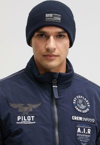 Navy beanie with a ribbed texture features a rectangular patch displaying brand details. Paired with a matching insulated jacket with embroidered accents.