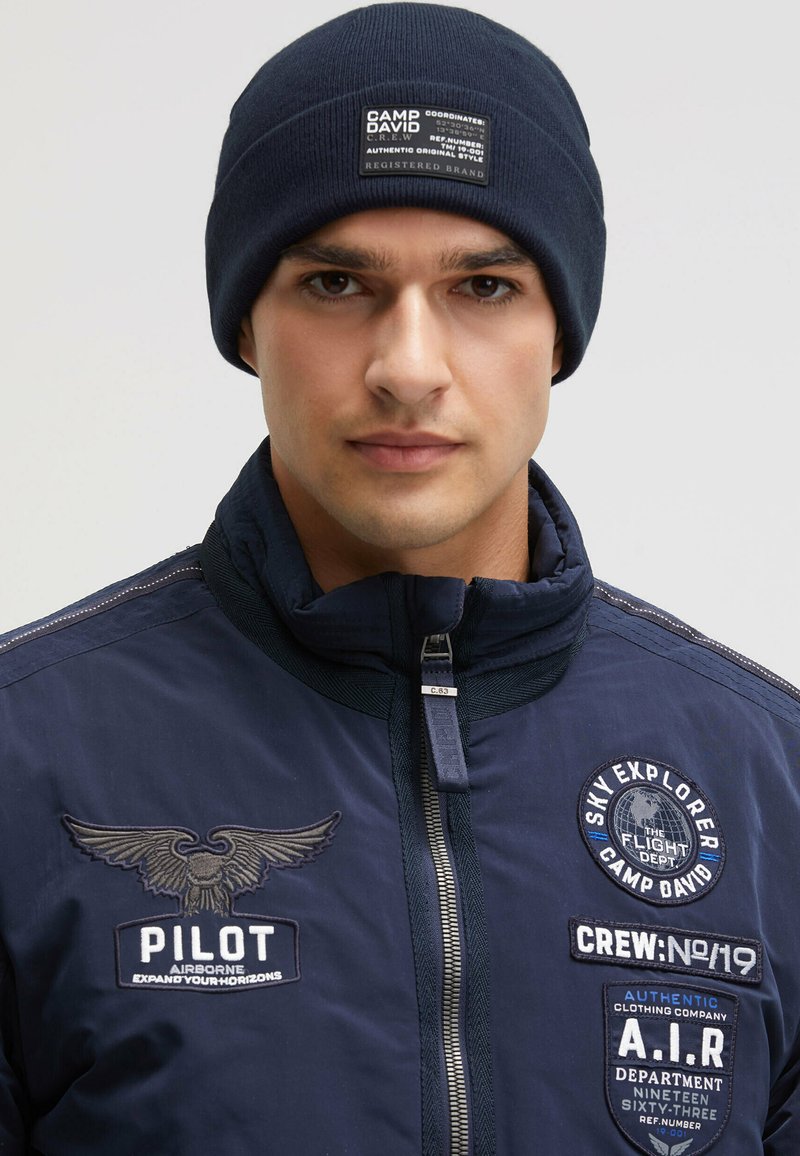 Navy beanie with a ribbed texture features a rectangular patch displaying brand details. Paired with a matching insulated jacket with embroidered accents.