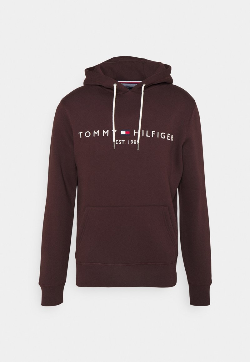 Sweat à capuche marron foncé avec poche avant, cordons blancs et logo "Tommy Hilfiger Est. 1985" en blanc avec des détails en rouge, blanc et bleu.