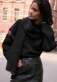 Schwarzer Strickpullover mit Kapuze, der über strukturierte Ärmel und ein Saumdetail verfügt. Kombiniert mit hoch taillierten schwarzen Lederhosen.