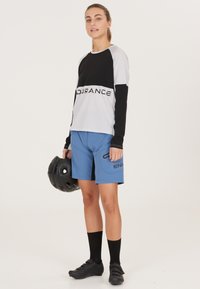 Langärmliges, schwarz-weißes Raglan-Shirt mit dem Schriftzug "ENDURANCE", kombiniert mit hellblauen Shorts mit schwarzen Akzenten und Taschen. Schwarzer Helm in der Hand.
